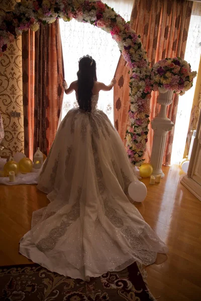 La mariée regarde par la fenêtre énorme dans la suite nuptiale révélant l'arrière de la robe de mariée. La face arrière d'un mariage blanc tient un cœur dans sa main . — Photo