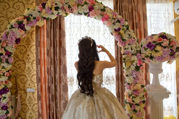 La sposa guarda fuori dalla finestra enorme nella suite nuziale rivelando il retro dell'abito da sposa. La parte posteriore di un matrimonio bianco tiene un cuore in mano . — Foto Stock