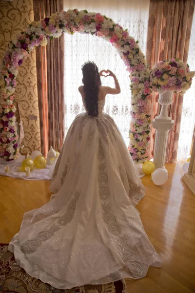 A noiva olha para fora da enorme janela na suite nupcial revelando a parte de trás do vestido de noiva. O lado de trás de um casamento branco tem um coração na mão . — Fotografia de Stock