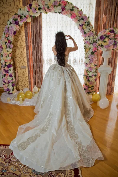 A noiva olha para fora da enorme janela na suite nupcial revelando a parte de trás do vestido de noiva. O lado de trás de um casamento branco tem um coração na mão . — Fotografia de Stock