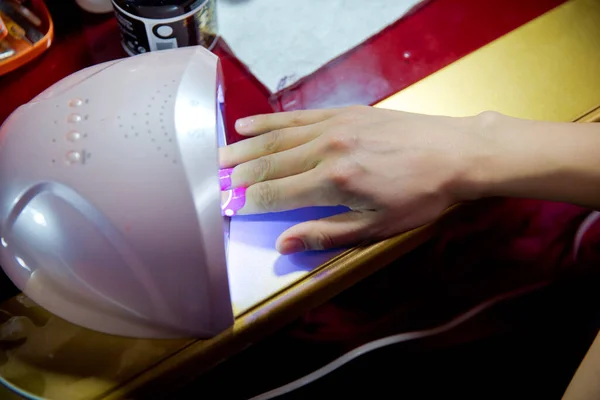 Mujer pone la mano en la lámpara ultravioleta led para curar la cubierta superior de esmalte de uñas. Fotografía horizontal en color. Vista de cerca de las manos femeninas con manicura de gel moderno rosa hermoso fresco . — Foto de Stock