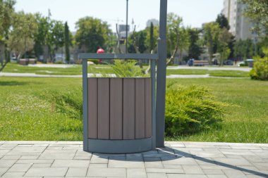 Parktaki ahşap bir muhafazanın içindeki kahverengi çöp tenekesi. Soldaki boşluğu kopyala. Parkta çöp kutusu var. Tahta bir vazo ve konforlu boş bir park bankı. .