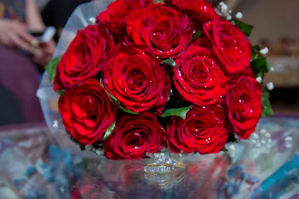 Glass table . Wedding rings next to a red flower bouquet selective focuse . Bride and groom with Engagement gold rings put on the table, and next to them lies a wedding bouquet .