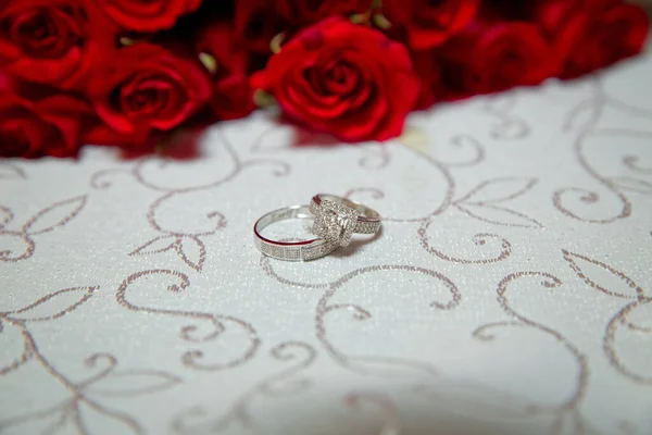 Mesa de vidro. Anéis de casamento ao lado de um buquê de flores vermelhas focuse seletivo. Noiva e noivo com anéis de ouro de noivado colocados sobre a mesa, e ao lado deles está um buquê de casamento . — Fotografia de Stock