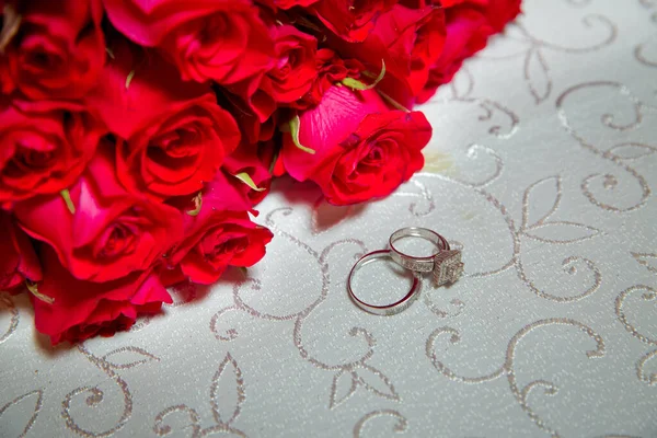 Tavolo di vetro. Anelli nuziali accanto a un mazzo di fiori rossi focuse selettivo. Sposa e sposo con anelli d'oro fidanzamento messi sul tavolo, e accanto a loro si trova un bouquet da sposa . — Foto Stock