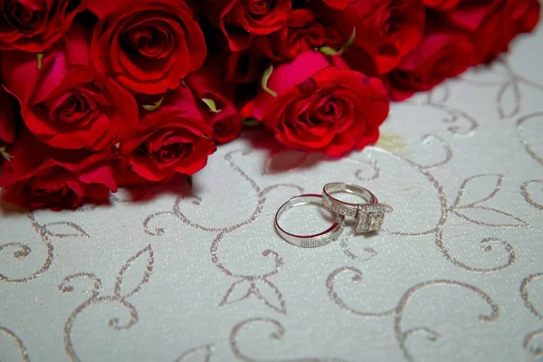 Tavolo di vetro. Anelli nuziali accanto a un mazzo di fiori rossi focuse selettivo. Sposa e sposo con anelli d'oro fidanzamento messi sul tavolo, e accanto a loro si trova un bouquet da sposa . — Foto Stock