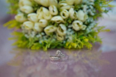 Alyanslar beyaz çiçek buketinin yanında seçici bir odak noktası. Nişan yüzükleriyle gelin ve damat masaya konmuş ve yanlarında bir düğün buketi var. Nişan yüzükleri.