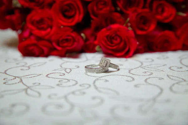 Mesa de vidro. Anéis de casamento ao lado de um buquê de flores vermelhas focuse seletivo. Noiva e noivo com anéis de ouro de noivado colocados sobre a mesa, e ao lado deles está um buquê de casamento . — Fotografia de Stock