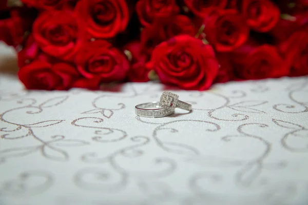 Tavolo di vetro. Anelli nuziali accanto a un mazzo di fiori rossi focuse selettivo. Sposa e sposo con anelli d'oro fidanzamento messi sul tavolo, e accanto a loro si trova un bouquet da sposa . — Foto Stock