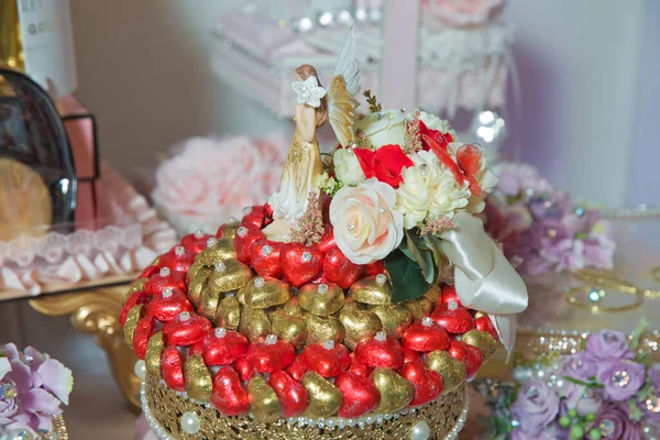 Basket with sweets stands on the table . The handle of the engagement basket . Heart-shaped candies in red and gold . The angel man on the basket . Engagement gold basket . — Stock Photo, Image