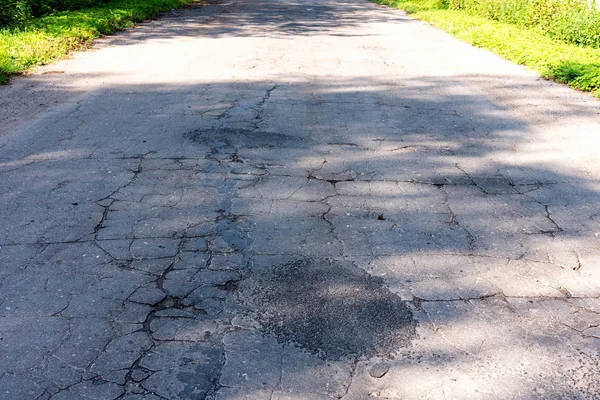 Broken empty asphalt country road surrounded by trees in sunny summer day. Road with holes in the green wood. A hole in the road.