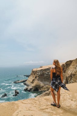 Yaz. Kız (sarışın) şort ve üst onu geri ile deniz kumlu bir uçurumun üzerinde duruyor. Peru, Paracas