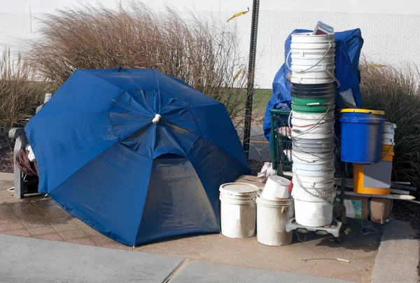 Homeless person`s belongings and buckets for can collection protected with umbrella.