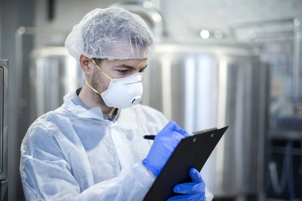 Technologist in white suit wearing hairnet and protective mask checking production in industrial factory.