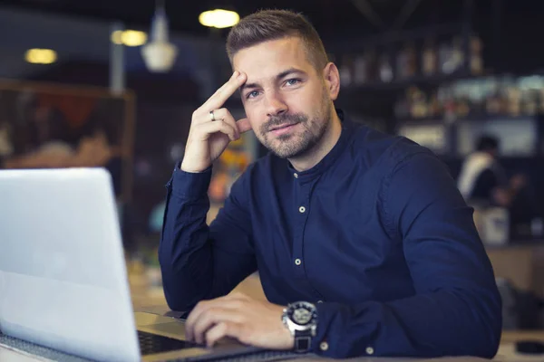 Ung kaukasisk affärsman sitter i cafeterian med bärbar dator på bordet. Smart kille tittar på kameran i caféet bar. — Stockfoto