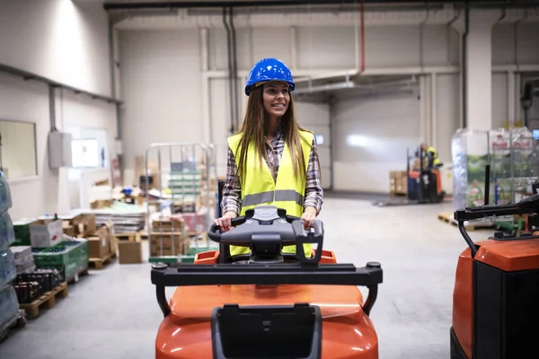 Vrouwelijke magazijnmedewerker met harde hoed en reflecterende veiligheidsuitrusting rijden heftruck machine in grote distributie magazijn centrum. Industriële fabrieksgebouw interieur. — Stockfoto