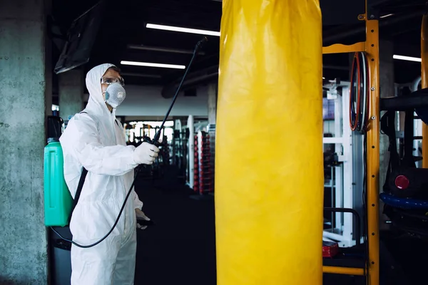 Gym disinfection and healthcare. Man in white protection suit disinfecting and spraying fitness equipment to stop spreading highly contagious coronavirus or COVID-19.