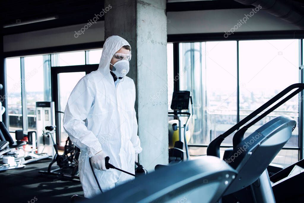 Gym disinfection of fitness equipment. Man in white protection suit disinfecting and spraying treadmill running tracks to stop spreading highly contagious coronavirus or COVID-19.