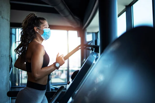 Spor kadın spor salonunda koşu bandında antrenman yapıyor ve küresel covid-19 virüsü salgınında kendini koronavirüsten korumak için maske takıyor..