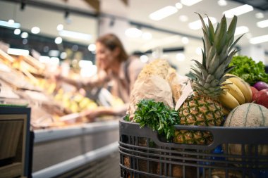Yiyecek, meyve ve sebze dolu süpermarketteki alışveriş arabasının kapanışı. Arka plandaki kadın ürünleri raflardan indiriyor..