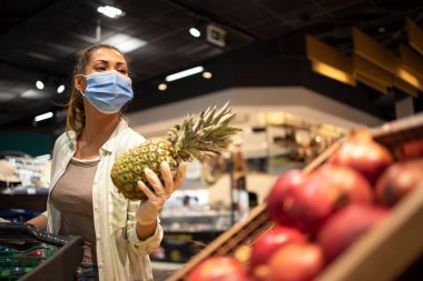 Hijyenik maskeli, lastik eldivenli ve markette alışveriş arabalı bir kadın covid-19 sırasında meyve alıyor ve salgın karantinasına hazırlanıyor..