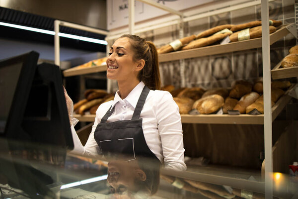 Female baker seller working on computer and selling bread in supermarket.