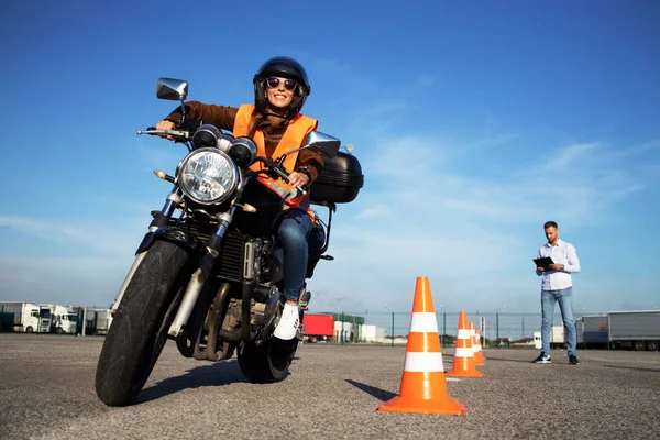 Kasklı bir kız öğrenci motosiklet dersleri alıyor ve pratik yapıyor. Arka plan trafik konileri ve kontrol listesi derecelendirme ve sürüş değerlendirmesi ile eğitmen. Motosiklet sürüş okulu.