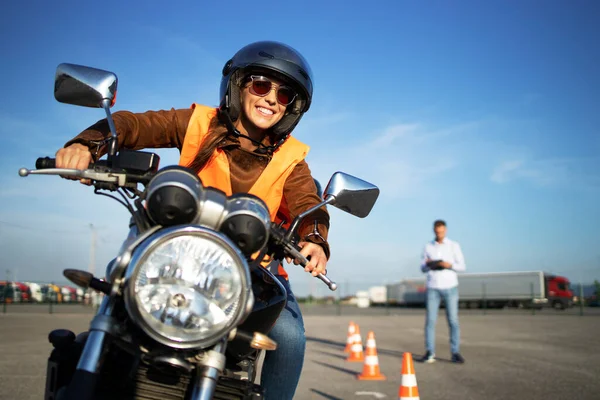 Kasklı bir kız öğrenci motosiklet dersleri alıyor ve pratik yapıyor. Arka plan trafik konileri ve kontrol listesi derecelendirme ve sürüş değerlendirmesi ile eğitmen. Motosiklet sürüş okulu.