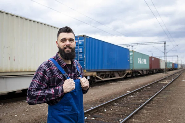 Railway worker proudly standing at train station. Shipping containers and freight train in background.