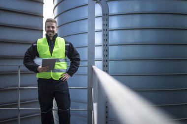 Endüstriyel depolama tankları arasında metal platformda duran ve kameraya bakan fabrika mühendisi portresi.
