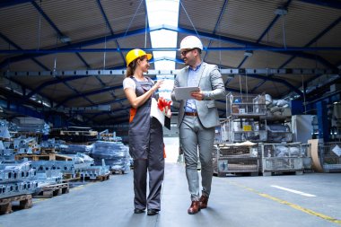 Üniformalı bir yönetici ve sanayi işçisi büyük bir metal fabrikasında yürüyor ve üretimin artmasından bahsediyor..
