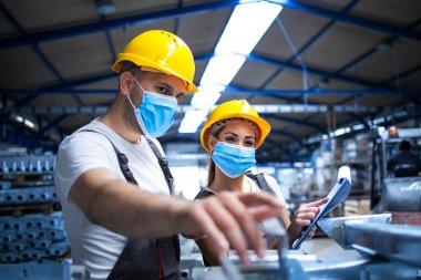 Yüz maskeli endüstriyel işçiler Corona virüsüne karşı korunuyor fabrikadaki metal parçalarını tartışıyorlar. COVID-19 salgını sırasında çalışanlar.