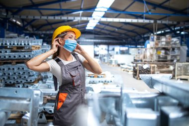 Üniformalı kadın fabrika işçisi ve endüstriyel üretim fabrikasında yüz maskesi takarken çekilmiş. COVID-19 salgını sırasında çalışanlar.