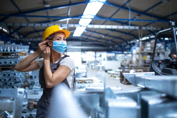 Üniformalı kadın fabrika işçisi ve endüstriyel üretim fabrikasında yüz maskesi takarken çekilmiş. COVID-19 salgını sırasında çalışanlar.