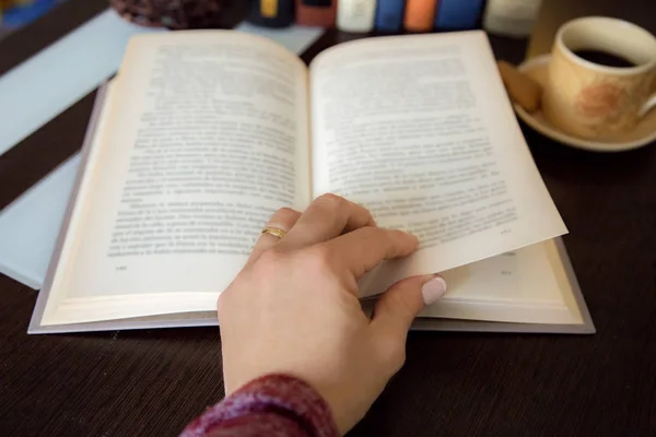 Detalhe da mão feminina girando uma página de livro em madeira escura com mais livros e xícara de café no fundo — Fotografia de Stock