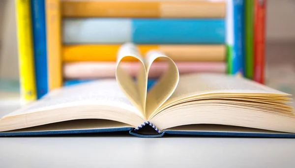 Ανοιχτό βιβλίο με δύο σελίδες διπλωμένα στο σχήμα της καρδιάς λεπτομέρεια και αρκετά πολύχρωμα βιβλία στο παρασκήνιο — Φωτογραφία Αρχείου