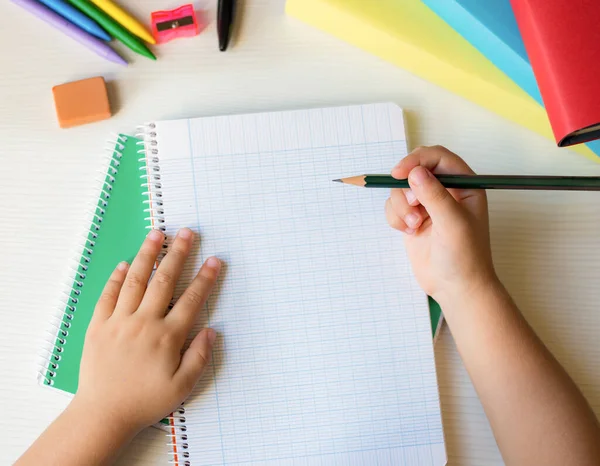 detail of children hands holding a pencil on a squared notebook ready to write on a white wooden desk with colorful school items