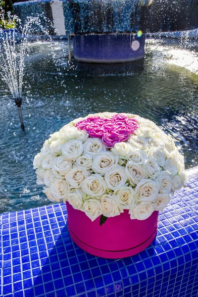 Bouquet of white roses with a heart of pink roses in a pink box near the fountain on a sunny day