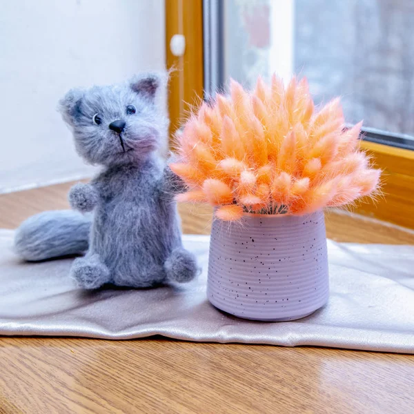 Funny gray knitted cat toy next to bouquet of fluffy dry spikelets on vases,  wooden window