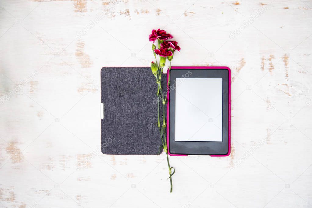 On the white worn background lies an open e-book, on it in the form of a bookmark is a maroon carnation flower.