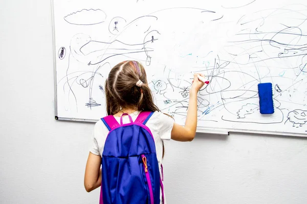 Ragazza con uno zaino blu sta con la schiena e disegna un pennarello su un consiglio scolastico bianco — Foto Stock