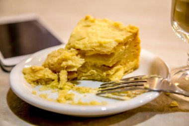 Bir tabak, bir cep telefonu ve hafif ahşap bir masa üzerinde cappuccino standı bir fincan Napolyon kek bir yarım yemiş dilim.