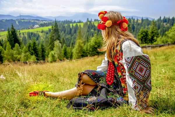 Chica en un viejo traje nacional ucraniano se sienta en un fondo de un paisaje verde de montaña en un día soleado — Foto de Stock