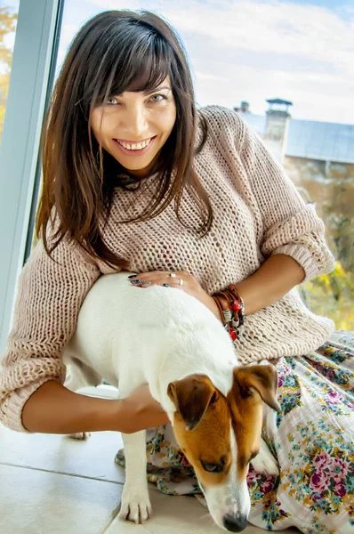 Mooie vrouw met donker haar tegen een raam zit op de vloer met een hondenras Jack Russell, herfststemming — Stockfoto