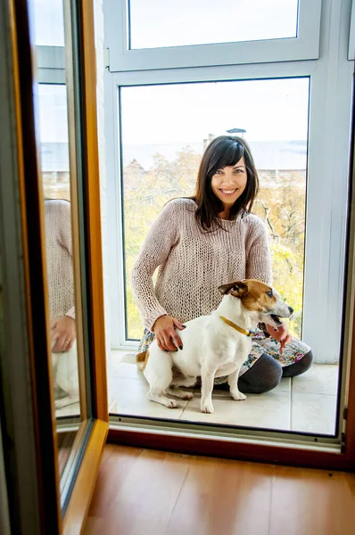 Mooie vrouw met donker haar tegen een raam zit op de vloer met een hondenras Jack Russell, herfststemming — Stockfoto