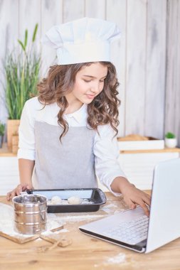 Kız mutfakta. Beyaz dizüstü bilgisayar. Çörek pişirme işlemi. Açık renkli.
