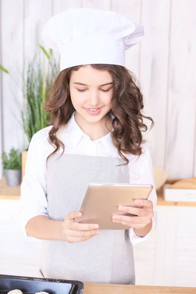 Girl with tablet in kitchen. Cooking buns. Light wooden background.