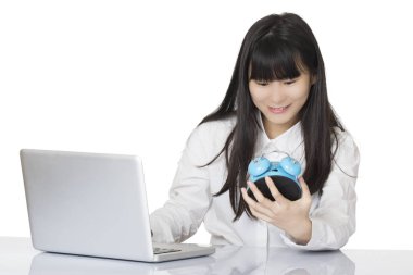 Beautiful Asian woman sitting at a desk very stressed out while holding a blue alarm clock isolated on a white background clipart