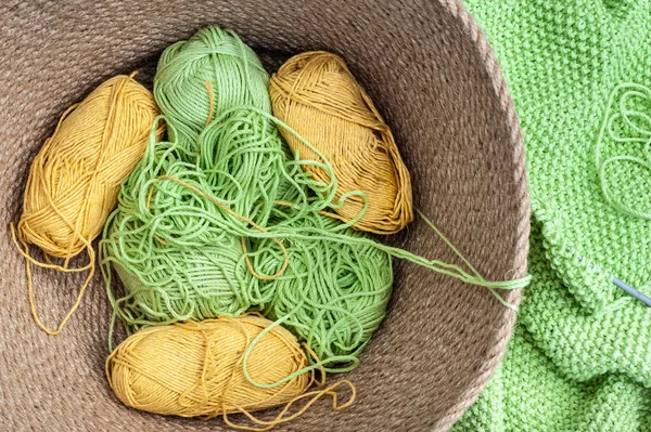 Basket with balls of green and yellow yarn close to green knitte