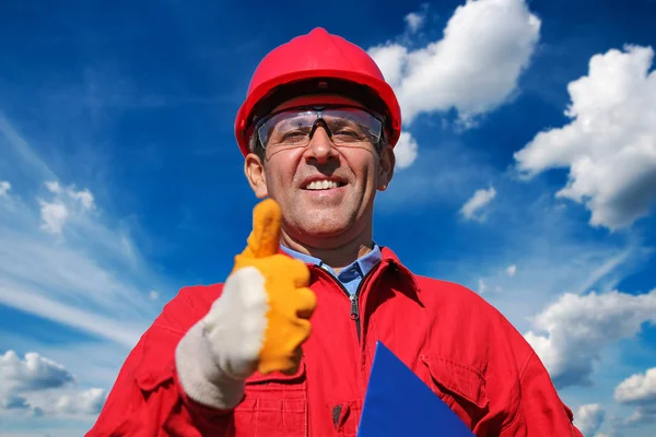 Worker in red hard hat and coveralls gesturing thumb up. Portrait of a smiling petrochemical engineer or construction supervisor.